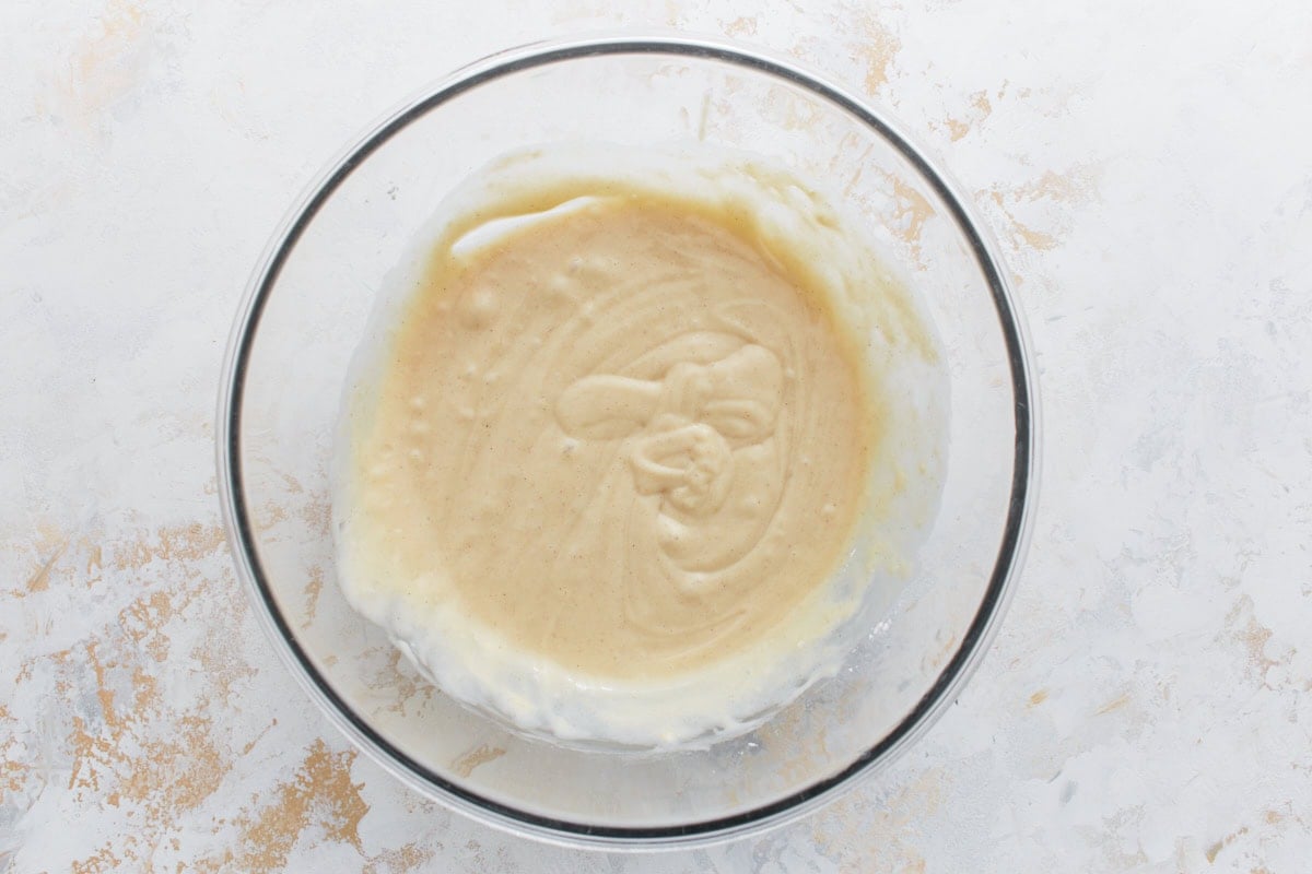 Glass mixing bowl filled with partially mixed cinnamon roll muffin batter on a light, textured background.
