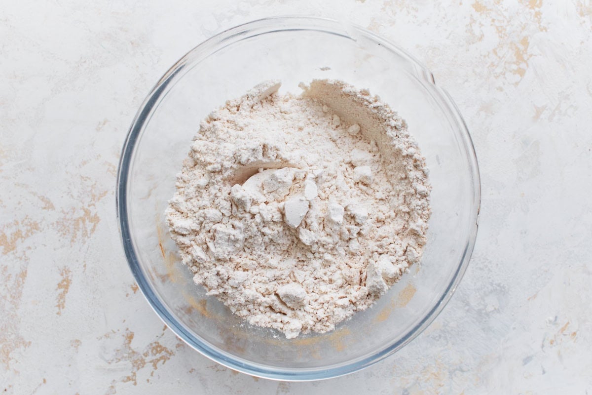 Overhead view of a clear glass bowl filled with mixed dry ingredients, including gluten-free flour and leavening agents, on a light textured surface.