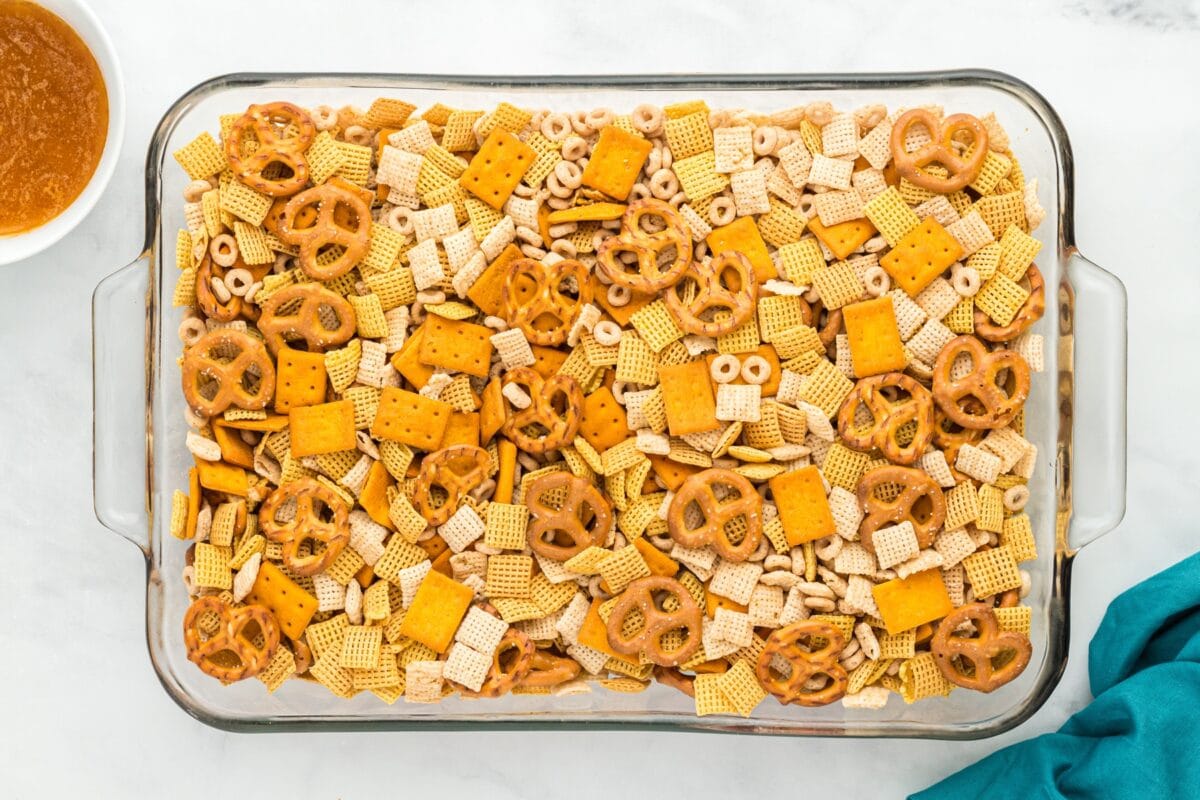 Glass baking dish filled with an unbaked mix of gluten-free cereals, pretzels, and crackers, with a bowl of sauce nearby.