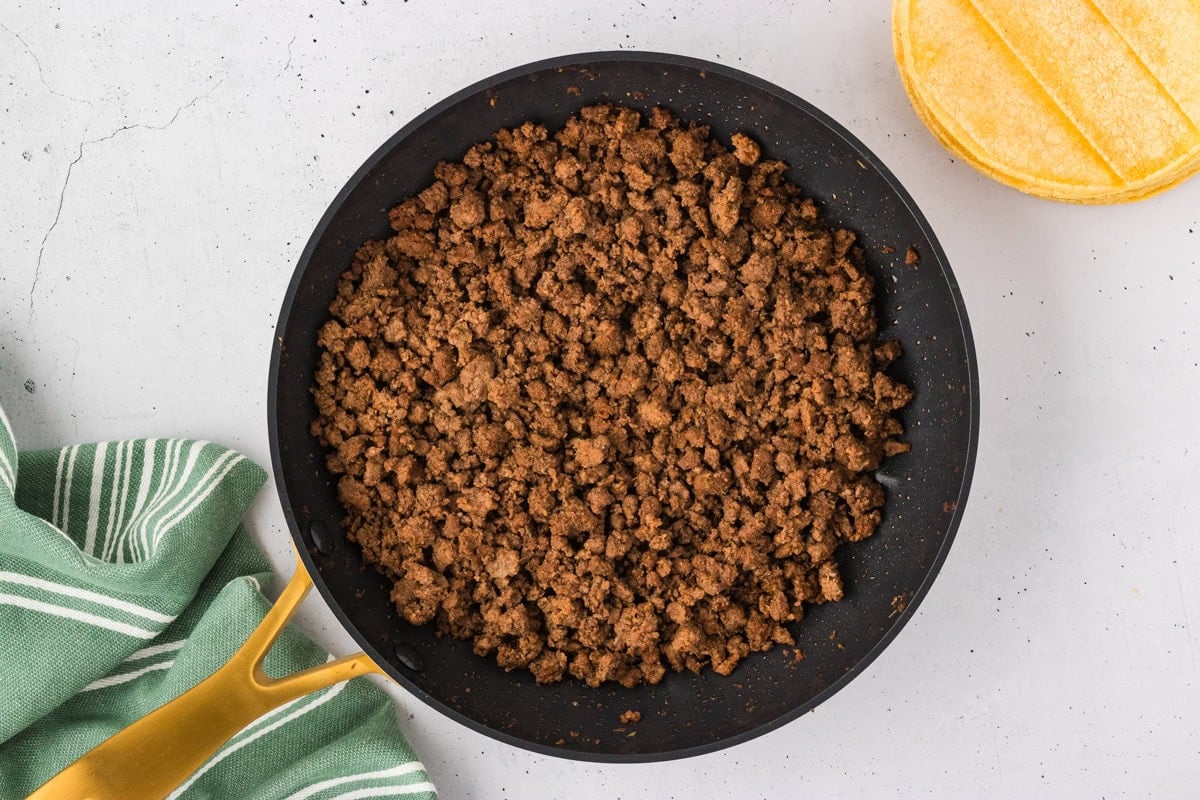 A skillet filled with cooked, seasoned ground beef next to a stack of corn tortillas and a green striped towel.