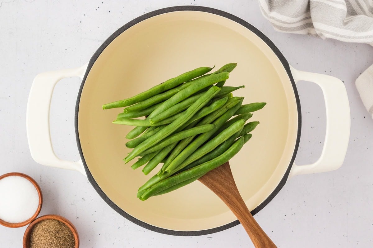 Raw green beans in a cream-colored pot being stirred with a wooden spoon, ready for blanching.