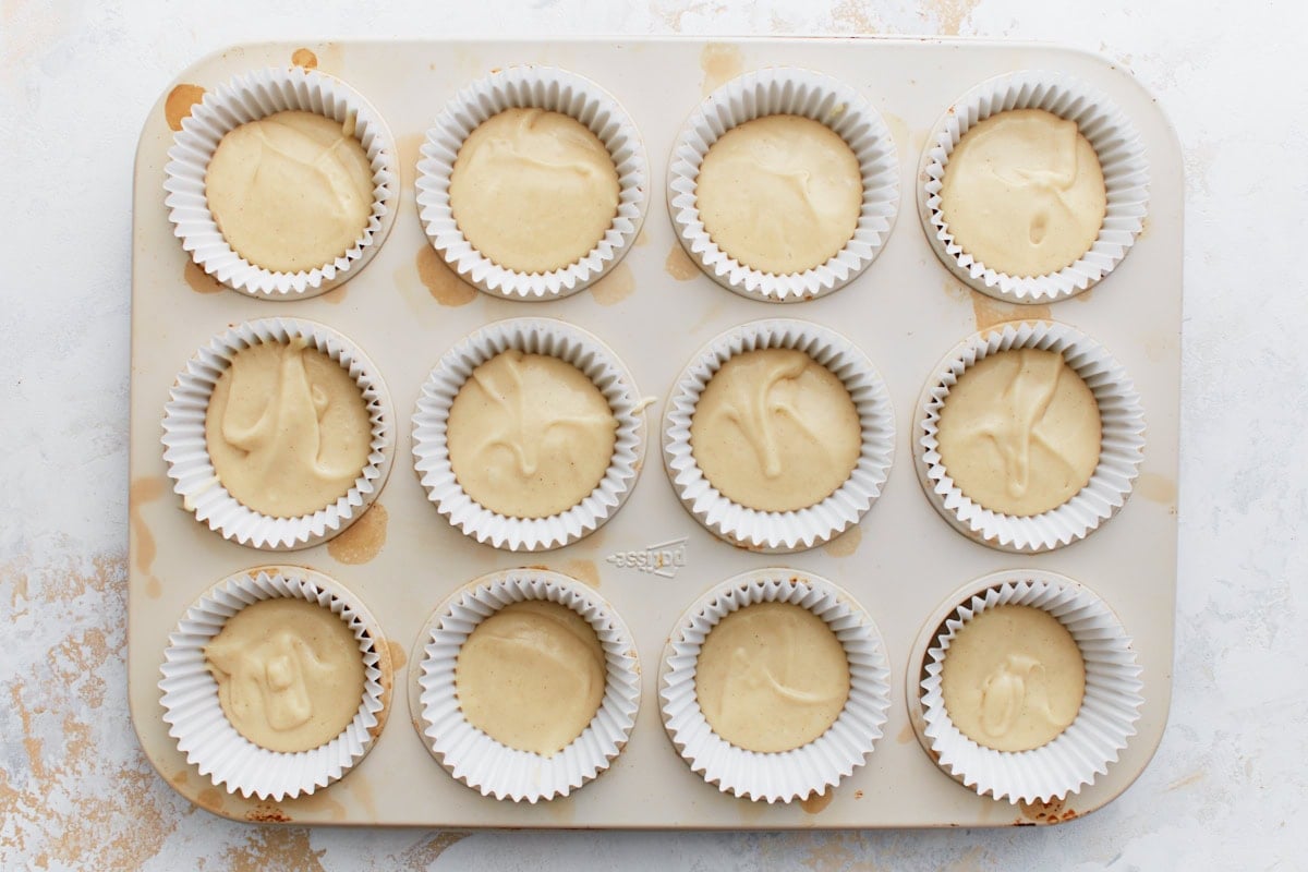 Muffin tin lined with white paper liners, each filled with thick gluten-free batter, ready for the cinnamon topping.