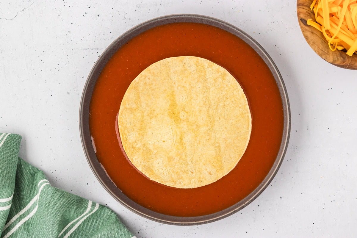 A corn tortilla soaking in a shallow bowl of red enchilada sauce with shredded cheese visible in a wooden bowl nearby.