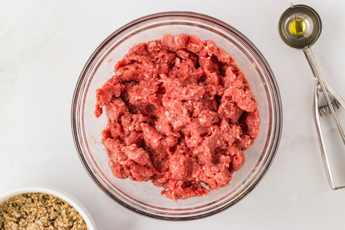 Raw ground beef in a glass mixing bowl with a cookie scoop and measuring spoon beside it.