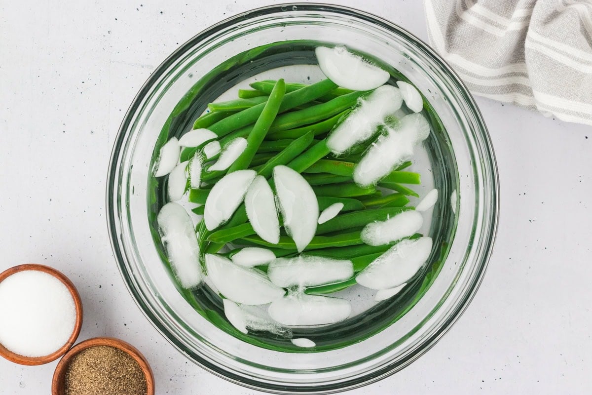 Blanched green beans in a clear bowl filled with ice water to stop cooking, with salt and pepper bowls nearby.
