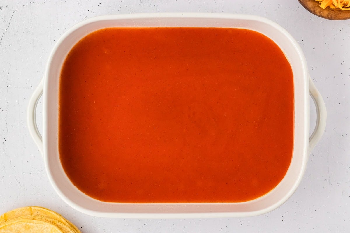 A 9x13 white baking dish with a layer of red enchilada sauce coating the bottom, ready for assembling enchiladas.
