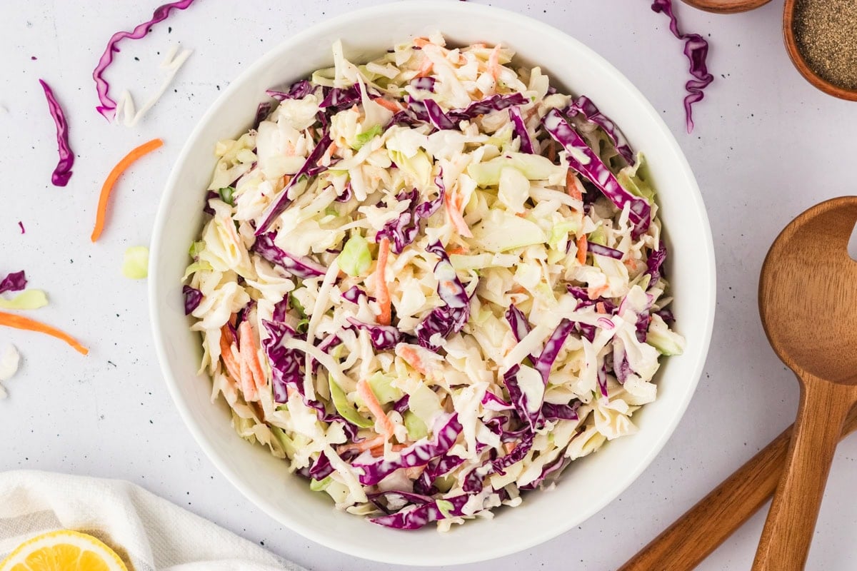 Overhead shot of finished coleslaw in a white serving bowl, surrounded by scattered bits of cabbage and carrot on a light background.