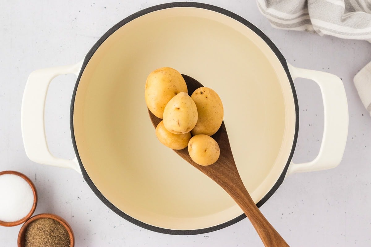 Golden baby potatoes in a cream-colored pot on a wooden spoon, ready to be boiled.