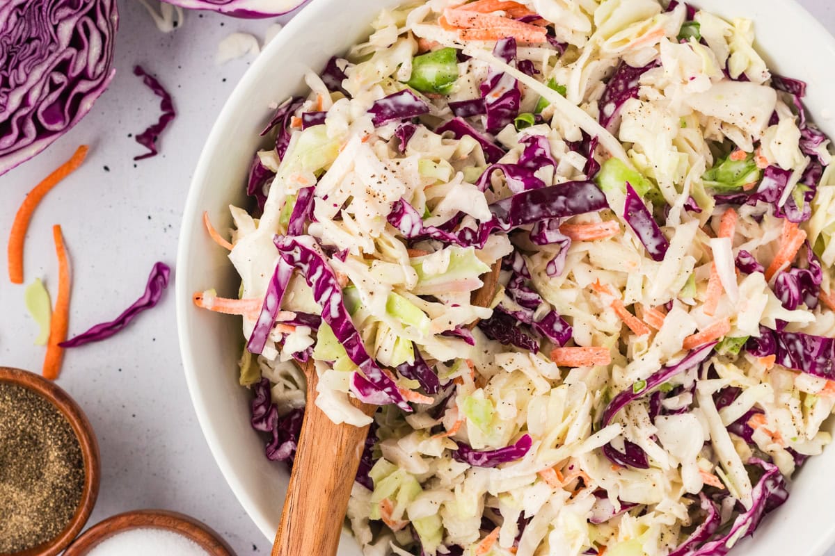 Close-up of coleslaw being scooped with wooden salad servers, showing the creamy texture and variety of vegetables.