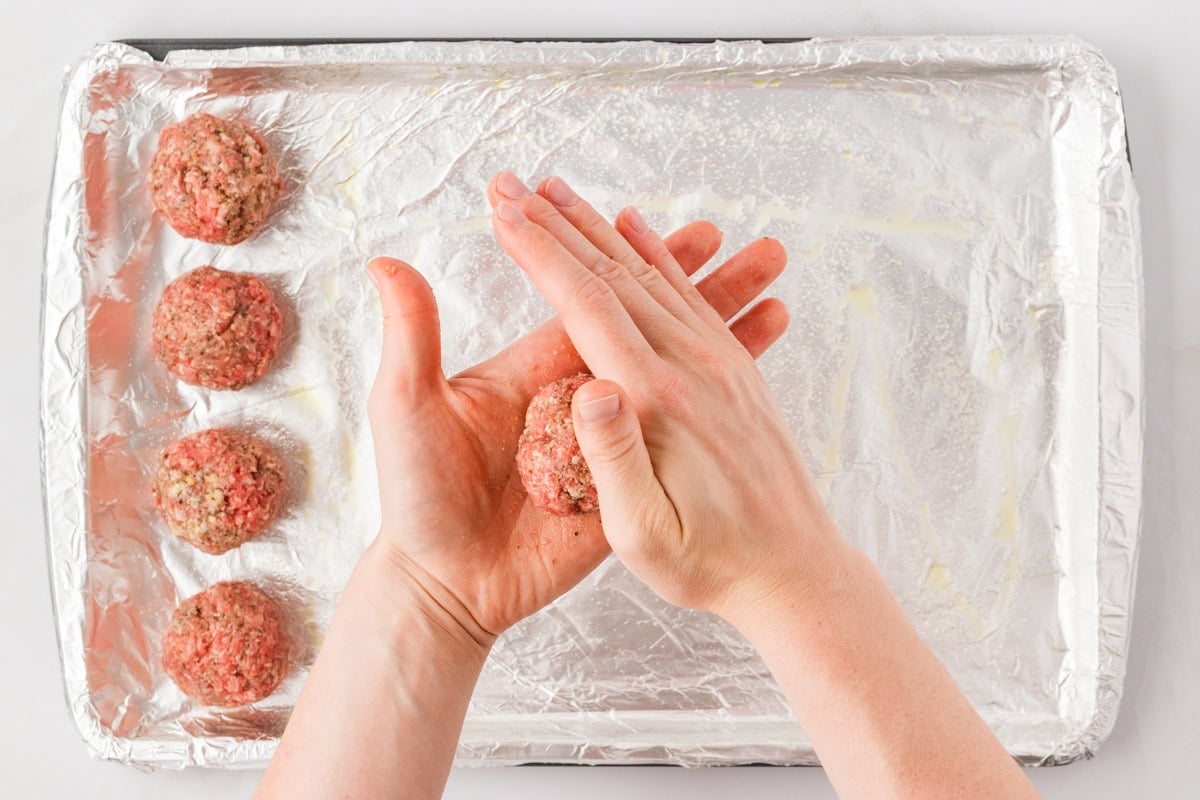 Hands shaping a meatball over a foil-lined baking sheet with already formed meatballs in the background.