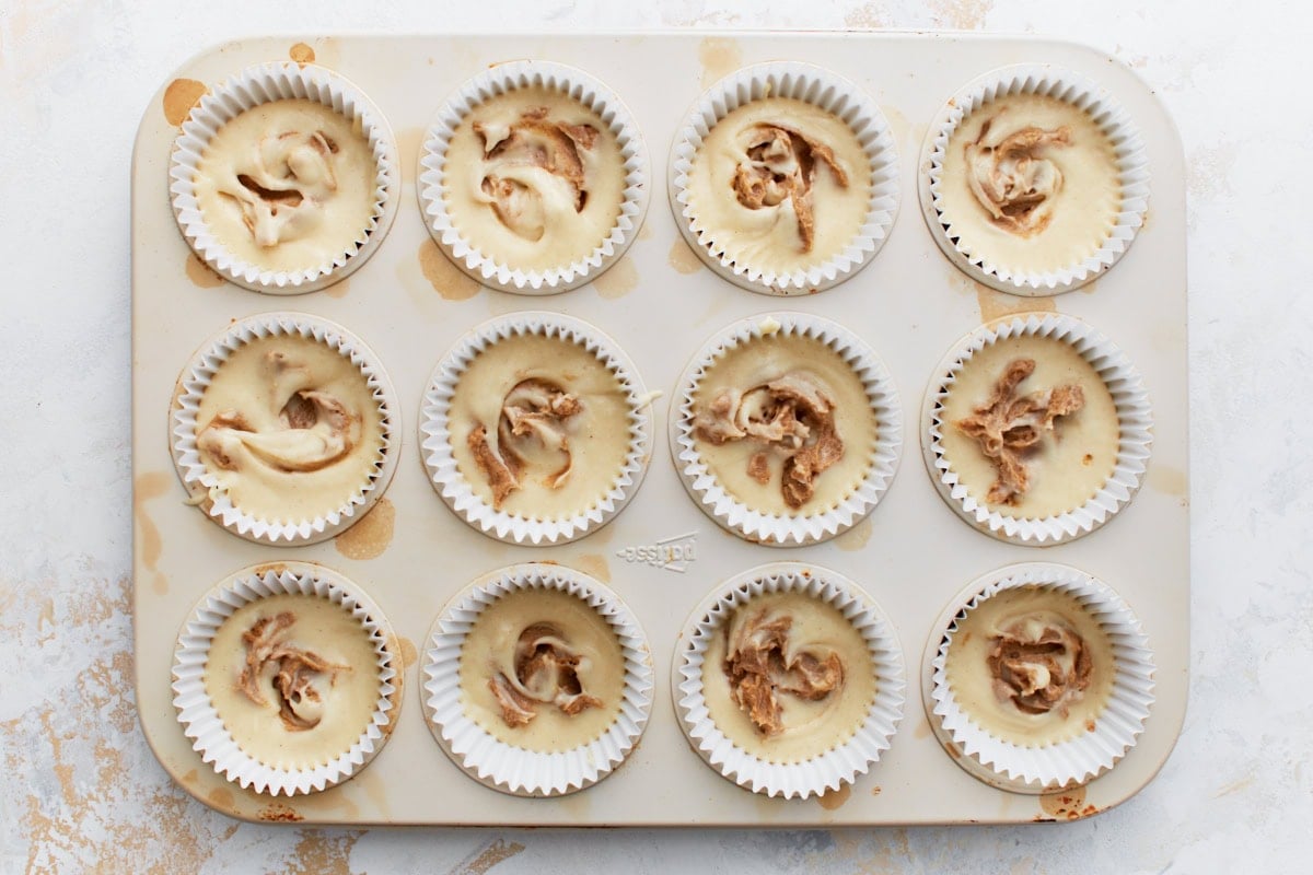 Muffin tin showing batter with cinnamon topping swirled into each cup, creating a marbled effect.