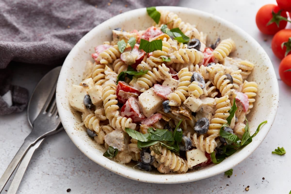 A side view of a bowl of creamy Italian pasta salad with a fork and spoon nearby, showcasing the texture and colorful ingredients.