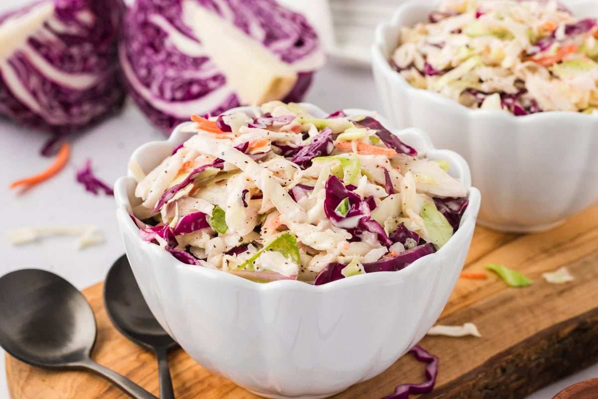 Two white bowls of finished dairy-free coleslaw on a wooden board, with a halved purple cabbage in the background and black serving spoons in front.