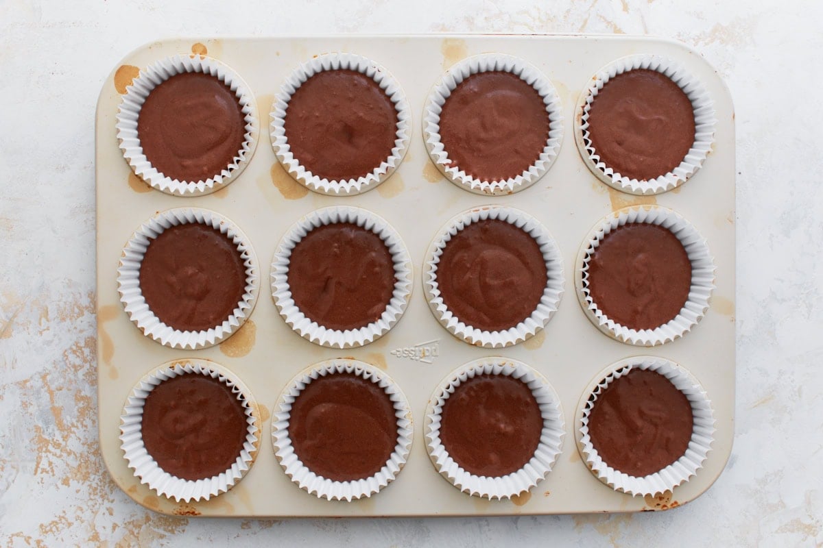 Muffin pan filled with white liners, each about two-thirds full of chocolate cupcake batter, ready to go into the oven.