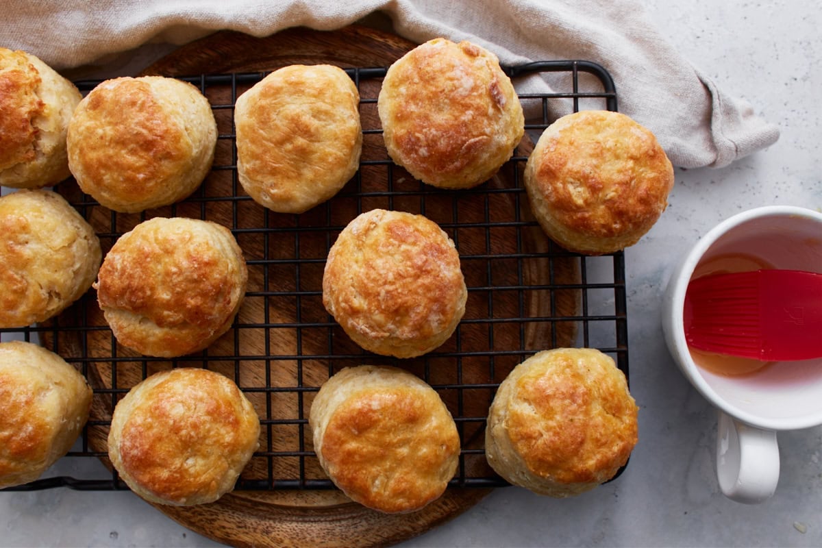 Baked biscuits cooling on a wire rack, with a pastry brush and cup of melted butter nearby.