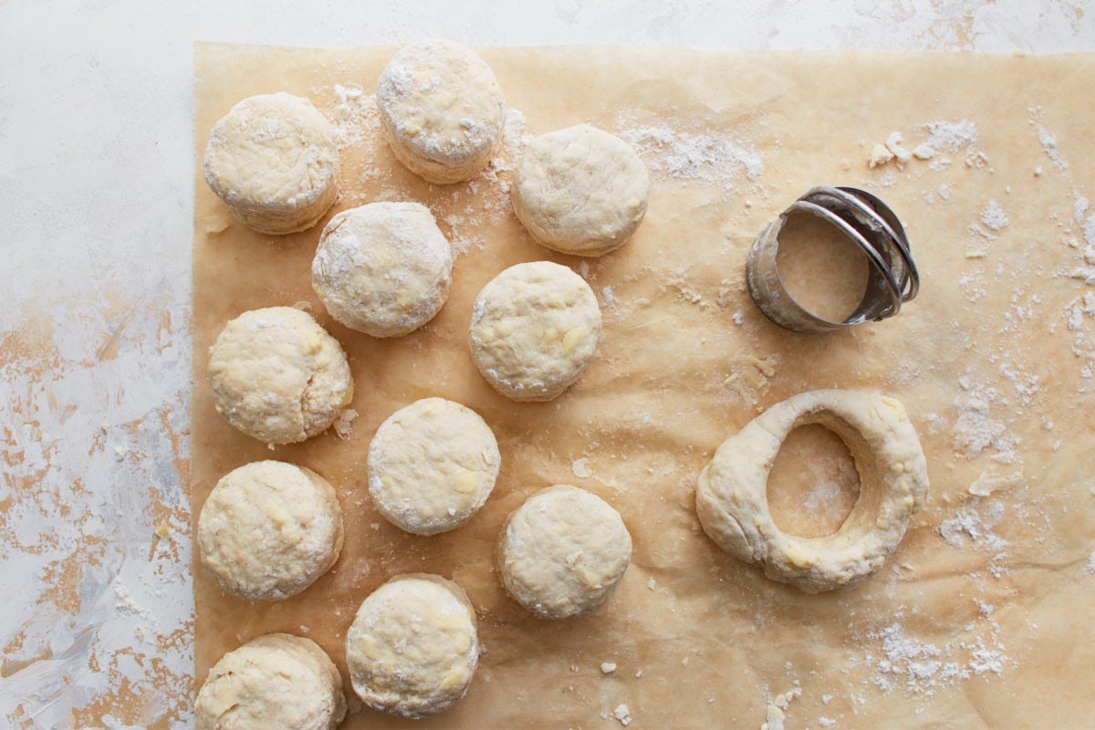 Unbaked gluten-free biscuit rounds on parchment paper with a biscuit cutter and scraps of dough nearby, ready to bake.