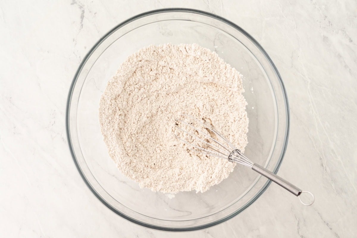 A large glass bowl with gluten-free flour mixture and a whisk on a white marble surface.