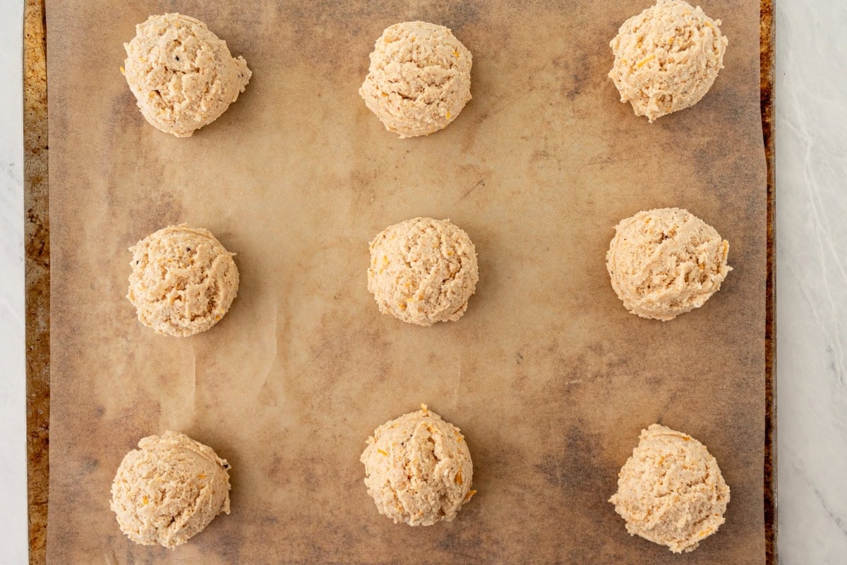 Nine scoops of biscuit dough evenly spaced on a parchment-lined baking sheet before baking.