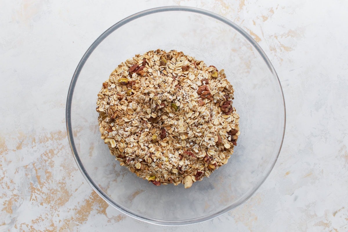 A large glass mixing bowl containing dry granola ingredients like gluten-free oats, chopped nuts, and seeds before any liquid has been added.