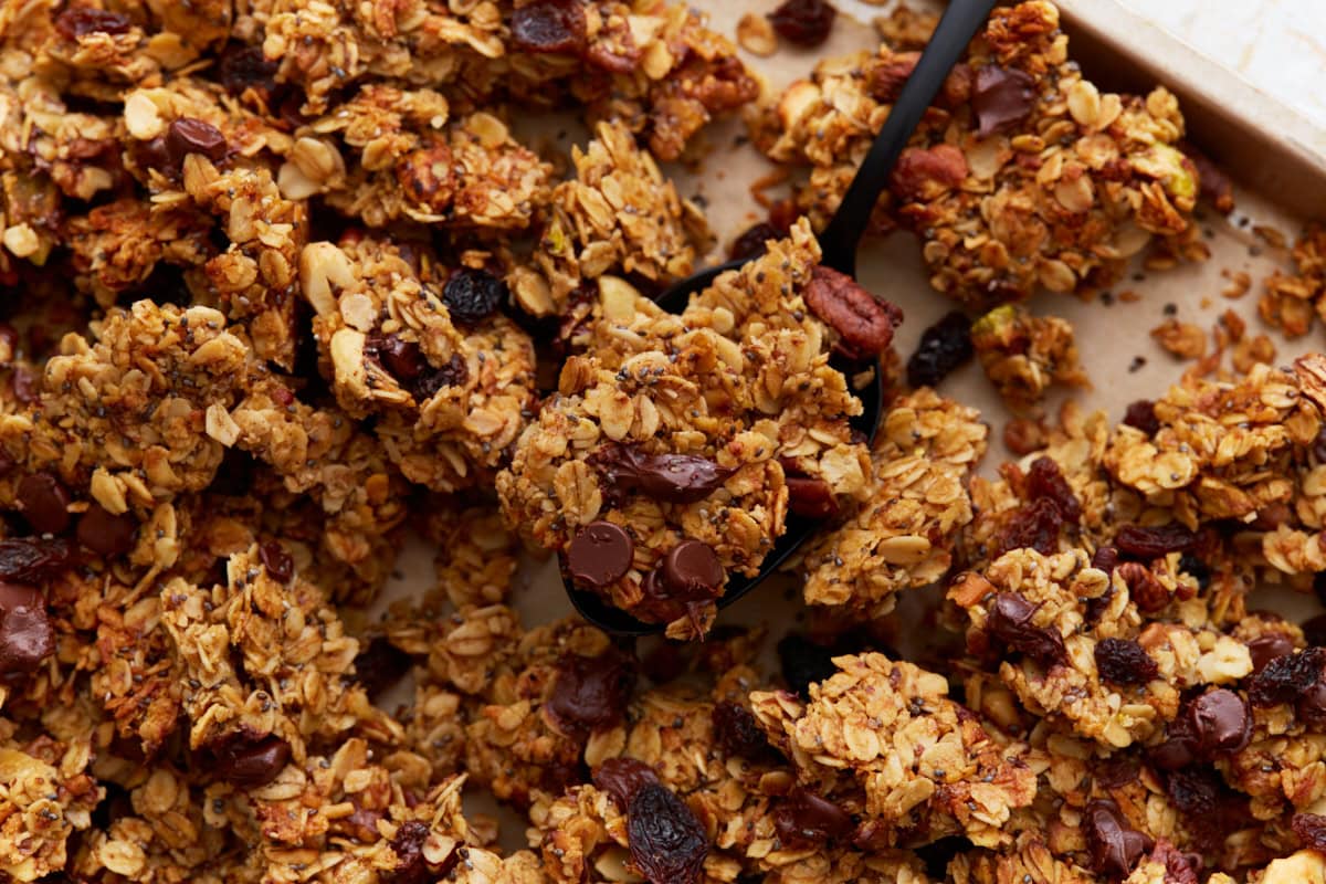 A close-up shot of a black spoon lifting a large granola cluster from the tray, highlighting the texture and visible mix-ins like raisins and chocolate chips.