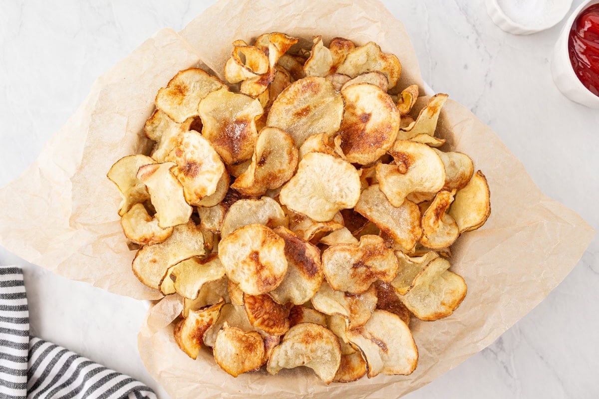 Finished batch of homemade gluten-free potato chips served in a parchment-lined bowl with ketchup on the side.