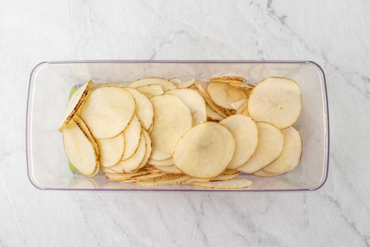 Thinly sliced raw potatoes arranged neatly in a clear container, ready for soaking.