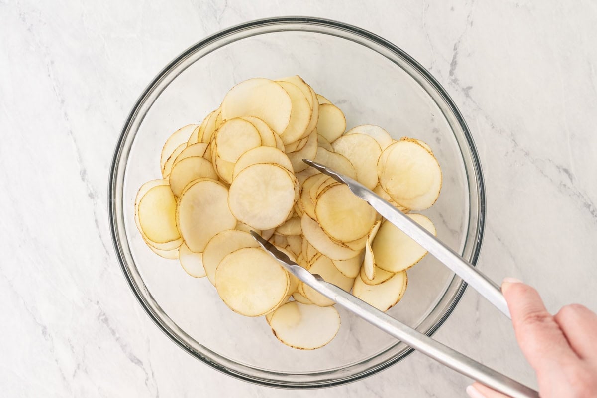 Potato slices in a glass bowl being tossed with tongs to coat with oil.