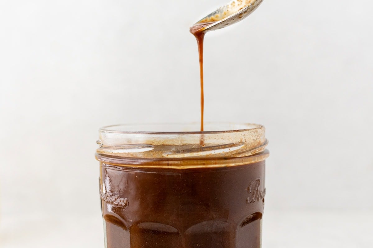 A close-up of a spoon dripping a thin stream of dark Worcestershire sauce back into the jar below.