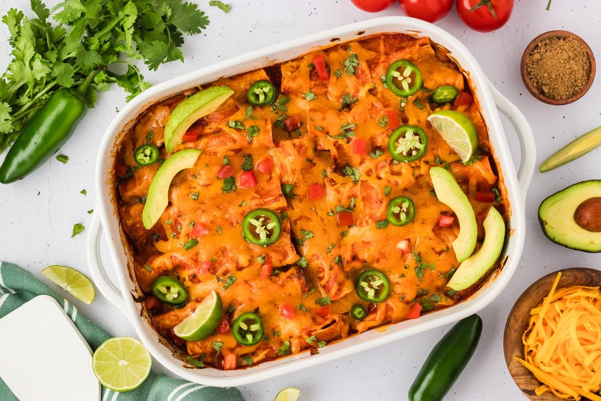 A finished dish of baked gluten-free beef and cheese enchiladas topped with melted cheese, jalapeños, tomatoes, avocado, lime wedges, and cilantro, surrounded by fresh ingredients.