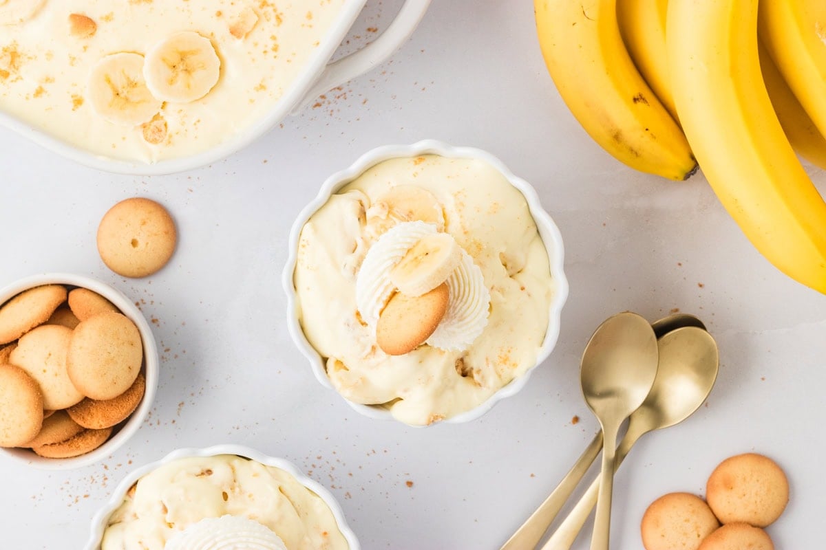 Flat lay of individual servings of gluten-free banana pudding in white bowls, garnished with whipped cream, banana slices, and cookies. Surrounded by extra cookies, fresh bananas, and gold spoons on a light background.
