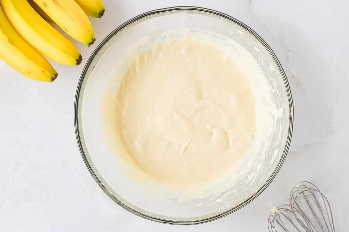 Creamy mixture of softened cream cheese and sweetened condensed milk in a glass mixing bowl, with bananas and a whisk in the background.