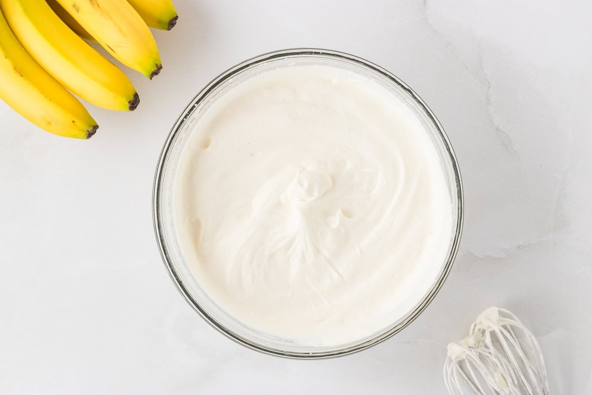 A bowl of freshly whipped cream with stiff peaks in a glass bowl on a white surface, bananas and a whisk nearby.