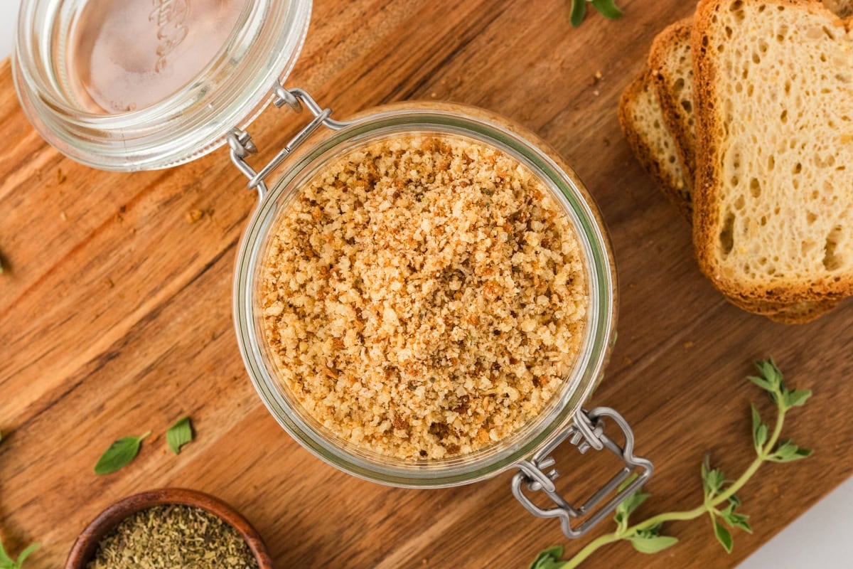Overhead view of a glass jar filled with gluten-free breadcrumbs, surrounded by fresh herbs and slices of gluten-free bread on a wooden board.