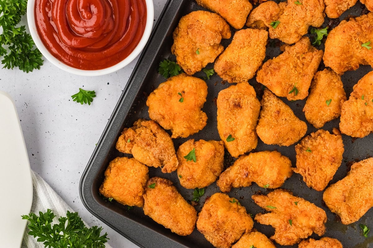 Overhead close-up of baked chicken nuggets on a sheet pan with a bowl of ketchup and sprigs of parsley nearby.