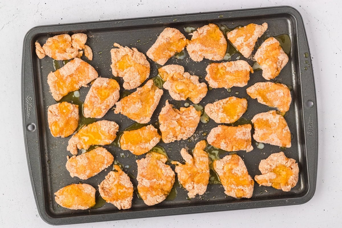 Breaded chicken nuggets arranged on a baking sheet, drizzled with melted butter, before baking.