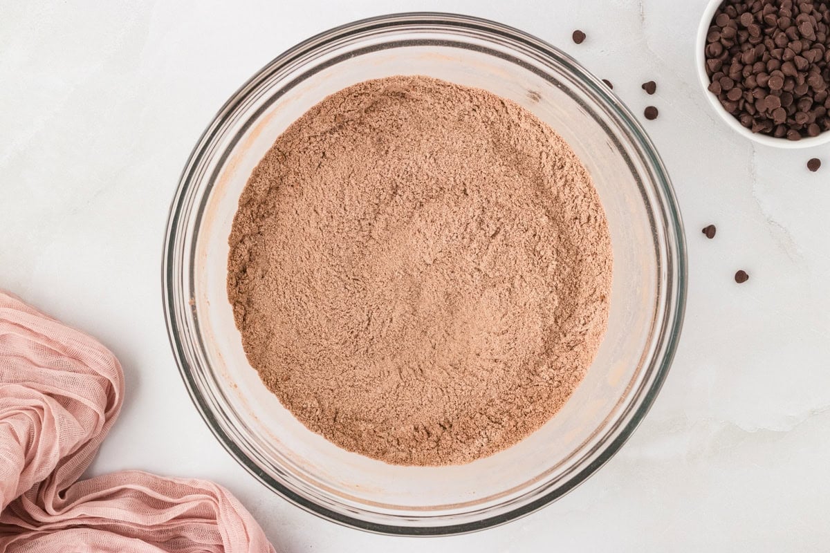 Glass bowl filled with blended dry ingredients for chocolate muffin batter, with chocolate chips and a pink cloth nearby.