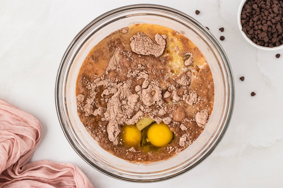 Glass mixing bowl with wet and dry muffin ingredients, including eggs, cocoa, and oil, before mixing.