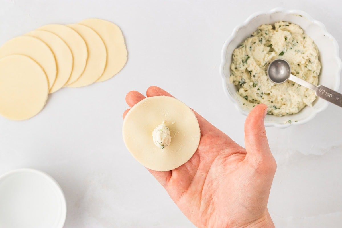 Hand holding a pasta round with a small spoonful of ricotta filling in the center, with more cut rounds and filling in the background