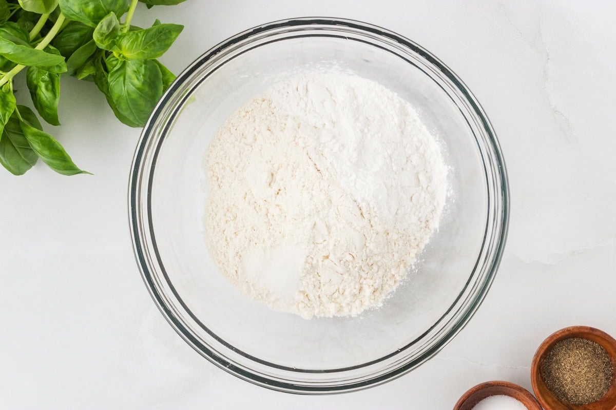 Glass bowl filled with gluten-free flour mixture, surrounded by fresh basil and small wooden bowls of salt and pepper