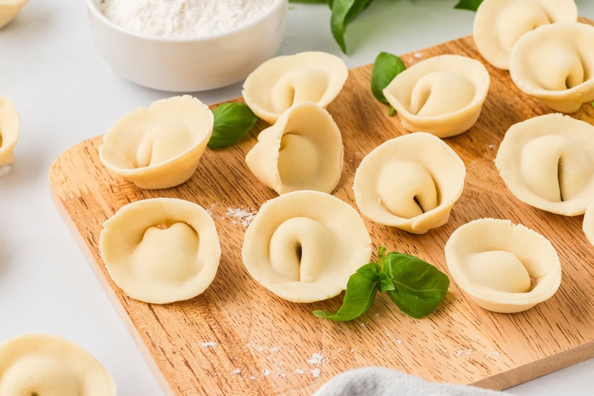 Wooden board topped with gluten-free tortellini and garnished with fresh basil leaves, flour bowl in background