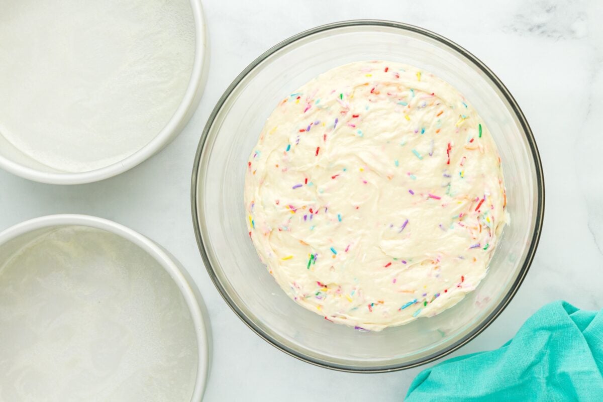 A bowl of thick gluten-free cake batter with colorful sprinkles folded in, next to prepared round cake pans.
