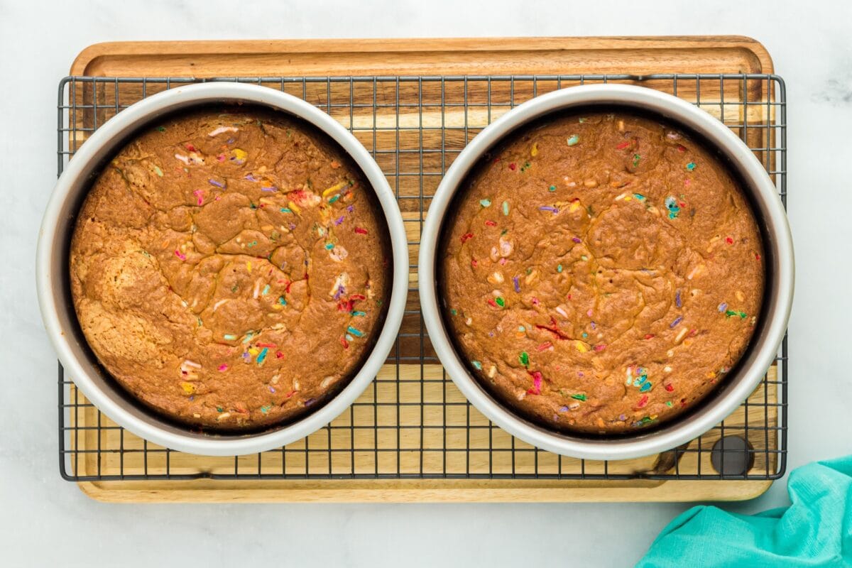 Overhead view of two baked funfetti cake layers cooling in their pans on a wire rack, with sprinkles visible on top.