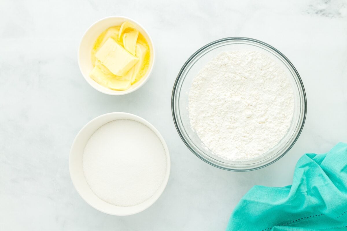 Overhead view of bowls containing butter, granulated sugar, and gluten-free flour on a white surface with a teal cloth.