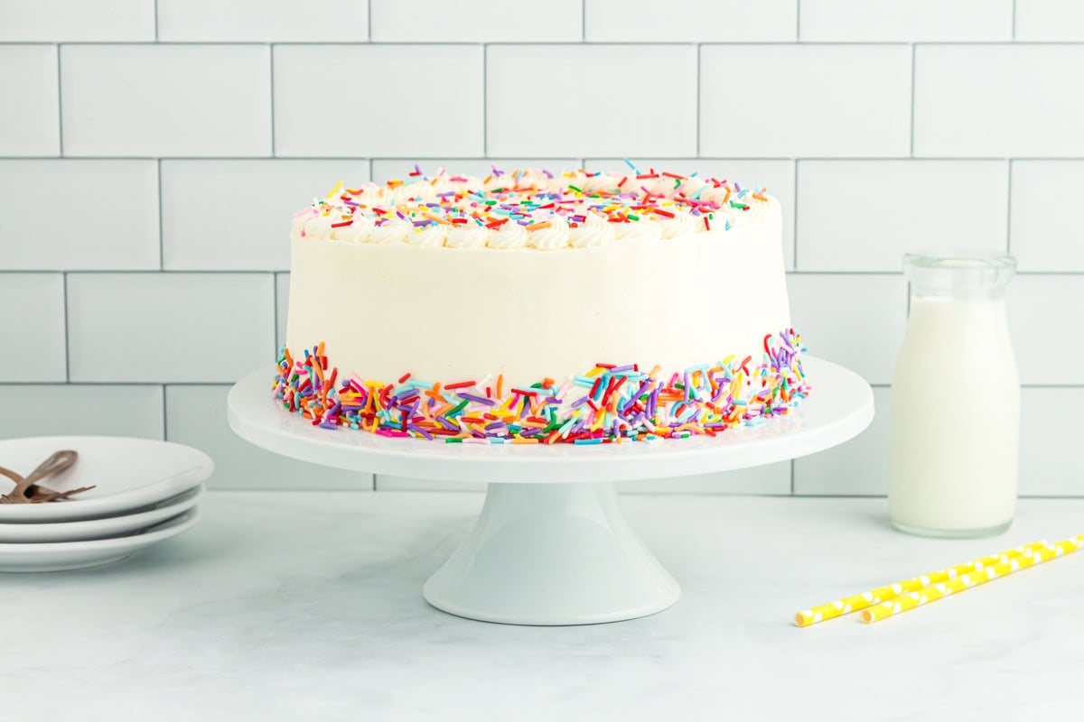 A fully frosted gluten-free funfetti cake on a white cake stand, decorated with rainbow sprinkles around the base and on top, with a bottle of milk and yellow straws nearby.