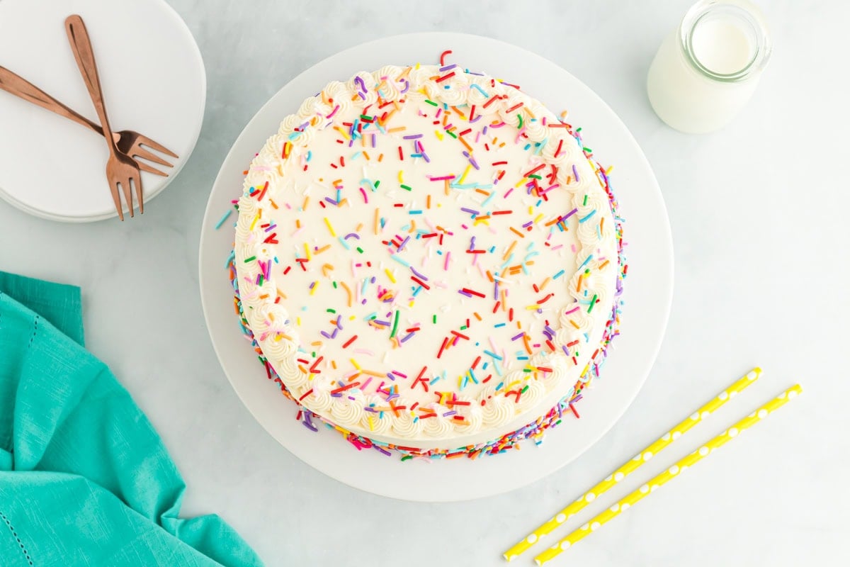 A top-down view of the finished funfetti cake, showing piped frosting swirls along the edge and a generous layer of rainbow sprinkles.