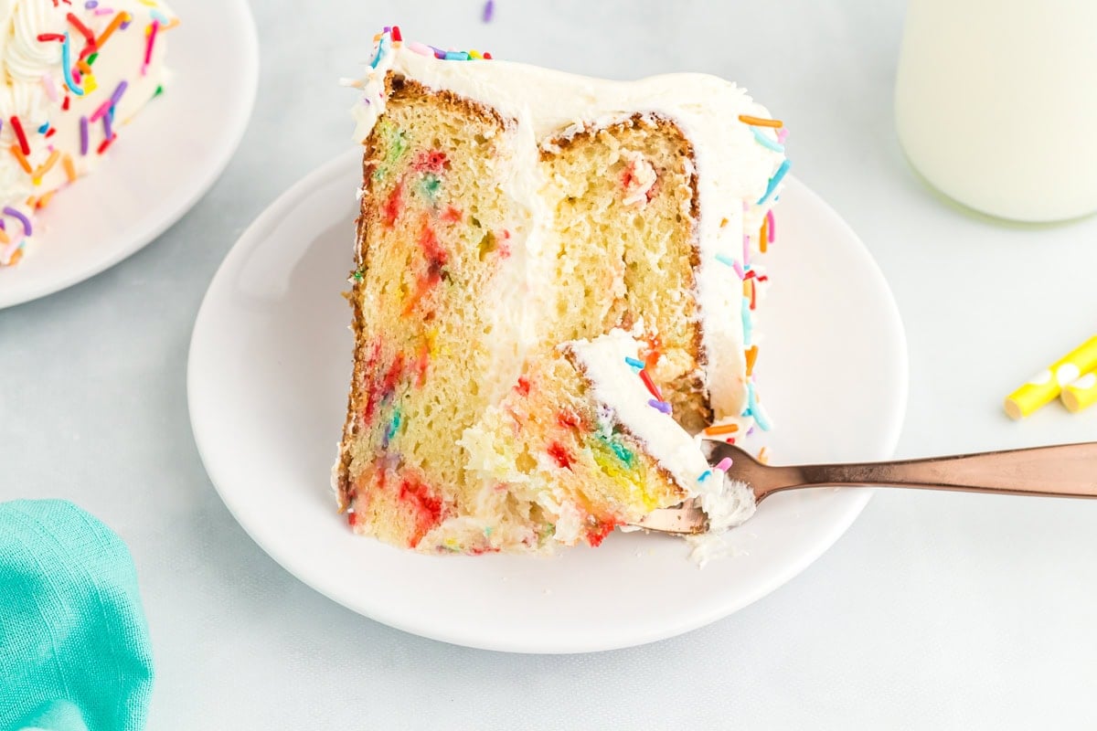 A slice of gluten-free funfetti cake on a white plate with a bite taken out using a copper fork, showing the soft texture and colorful sprinkles inside.