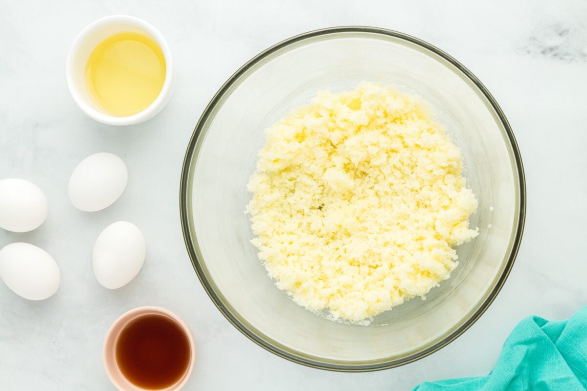 A mixing bowl with creamed butter and sugar surrounded by eggs, vanilla extract, and oil, ready to be combined.