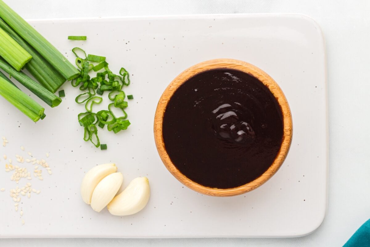 Bowl of homemade hoisin sauce on a white platter with sliced green onions, garlic cloves, and sesame seeds.