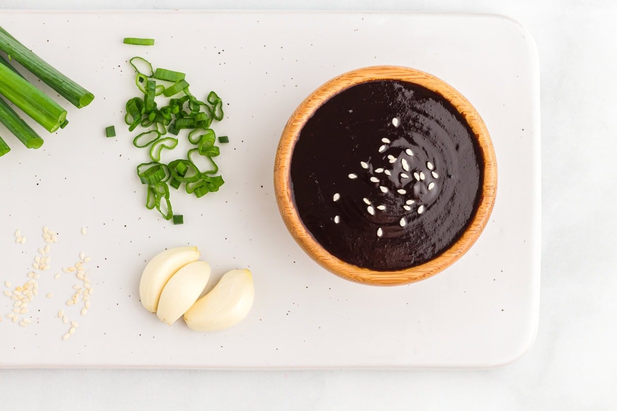 Overhead view of a bowl of hoisin sauce topped with sesame seeds, surrounded by fresh green onions, garlic cloves, and sesame seeds on a white background.