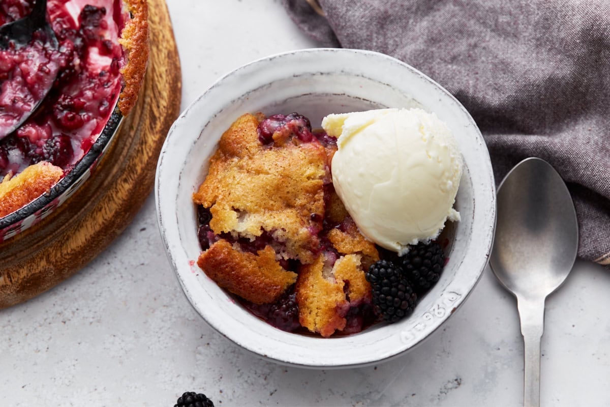 Serving of warm blackberry cobbler in a white bowl, topped with a scoop of vanilla ice cream and garnished with fresh blackberries.